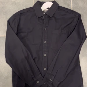 Svart overshirt från Jack & Jones - Stilren svart overshirt jacka från Jack & Jones med klassisk krage och bröstficka. Den har långa ärmar och knappar hela vägen framtill. Perfekt för dig som gillar enkel och clean stil. Materialet är mjuk bomull som känns skönt mot huden. Den är knappt använd o storleken är S