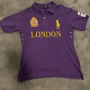 Lila Polo Ralph Lauren t-shirt London - Snygg lila t-shirt från Polo Ralph Lauren med klassisk krage, korta ärmar och broderad gul logga samt texten 'LONDON' på bröstet. Stor vit siffra 2 på ryggen och ärmen. Tillverkad i mjuk bomull, perfekt för dig som gillar sportig stil.