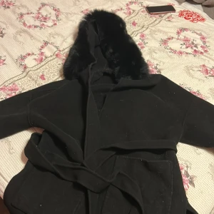 Svart kappa med fuskpäls och huva - Snygg svart kappa med stor huva och mjuk fuskpäls runt kanten. Jackan har bälte i midjan för en schysst siluett och är perfekt för kyliga dagar. Klassisk och stilren design som funkar till många outfits.