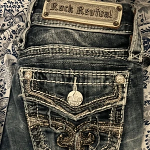 Rock Revival bootcut jeans  - Snygga blå bootcut jeans från Rock Revival med låg midja. Jeansen har detaljerade broderier och dekorativa knappar på bakfickorna. Typ storlek 24-25 och passar mig som är ca 162 perfekt i längd