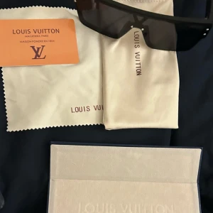 Svarta solglasögon  - Säljer ett par svarta, rektangulära solglasögon med bred båge och mörka linser. Glasögonen har en modern, kantig design och kommer med originalfodral, putsduk och certifikat. Perfekt accessoar för en trendig look.