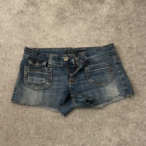 Blå jeansshorts från ONLY - Snygga blå jeansshorts från ONLY med slitna detaljer och rå kant nedtill. Shortsen har flera fickor fram och bak, samt coola metallknappar och en D-ring i midjan. Passformen är lågmidjad och materialet är klassisk denim. Perfekta för sommarens alla äventyr!