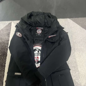 Svart parkajacka Geographical Norway - Svart parkajacka från Geographical Norway med huva, flera praktiska fickor och broderade loggor på bröstet och ärmen. Jackan har dragkedja framtill och är fodrad för extra värme. Perfekt för kalla dagar och snygg streetstil.