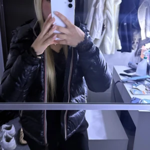 Svart Moncler pufferjacka med huva - Säljer en svart pufferjacka från Moncler med huva och klassisk logga på ärmen. Jackan har dragkedja framtill med snygga rödvita detaljer längs kanten. Perfekt för kalla dagar och riktigt skön att bära.