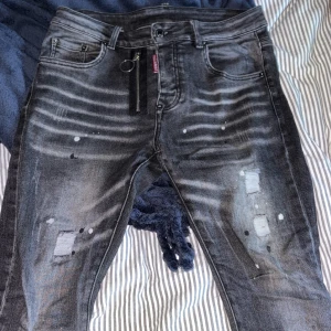 Svarta Dsquared2 jeans med slitningar - Säljer ett par svarta Dsquared2 jeans med coola slitningar, blekta detaljer och färgstänk. Jeansen har dragkedjedetalj framtill och klassisk femficksdesign. Märkespatch bak i svart läder och röd Dsquared2-tag vid fickan. Tillverkade i Italien. Ända felet är att metallen på läderlappen backtill är trasig, men går att limma.
