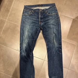 Blå jeans från Nudie Jeans Co - Snygga blå jeans från Nudie Jeans Co med klassisk femficksdesign och kontrastsömmar. Jeansen har en rak passform och coola slitningar framtill samt Nudie’s ikoniska broderi på bakfickorna. Perfekta för en avslappnad och trendig look.