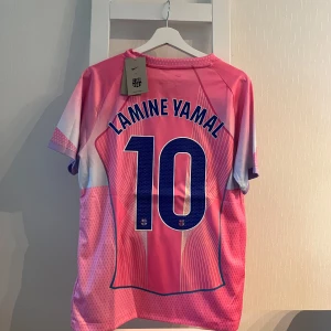 FC Barcelona Lamine Yamal rosa tröja Nike - Säljer en FC Barcelona fotbollströja med Lamine Yamal och nummer 10 på ryggen. Tröjan är rosa med vita och blå detaljer, har korta ärmar och är tillverkad i ett lätt, ventilerande material. Nike-logga, Barca-emblem och Spotify-logga framtill.