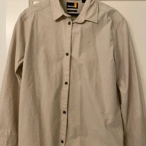 Beige skjorta från Timberland - Stilren beige skjorta från Timberland med relaxed fit. Skjortan har klassisk krage, svarta knappar och broderad logga på bröstet. Tillverkad i bomull, perfekt för dig som gillar en enkel och clean look.