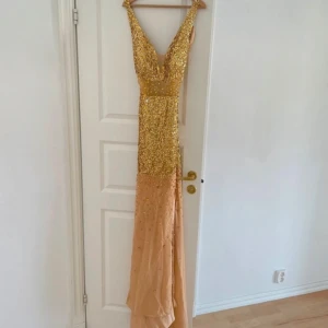 Glittrig långklänning i guld och champagne - Säljer en maxad långklänning med djup v-ringning, täckt av guldiga paljetter och stenar upptill och överdelen. Nederdelen är i champagnefärgad, tunn mesh med glittrande detaljer. Klänningen är ärmlös och har en figurnära passform som ger en riktigt glam känsla.