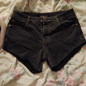 Svarta jeansshorts från In Blue - Svarta jeansshorts från In Blue med rå kant nertill och klassiska fem fickor. Shortsen har kontrastsömmar i gult och en snygg passform med låg midja. Perfekta för varma dagar och en avslappnad stil.