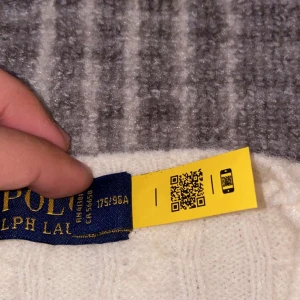 Vit kabelstickad tröja Polo Ralph Lauren - Kabelstickad vit tröja från Polo Ralph Lauren med klassisk rund halsringning och broderad marinblå logga på bröstet. Tröjan har långa ärmar och ribbade muddar för en snygg passform. Perfekt för dig som gillar stilrena och tidlösa plagg.