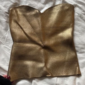 Guldig tubtopp från Zara - Säljer en snygg guldig tubtopp från Zara i storlek S. Toppen är axelbandslös med en glansig metallic-finish och har en lätt strukturerad yta. Perfekt för att sticka ut med en cool look. Stängs med en diskret dragkedja i sidan. Köpt på här på Plick men i ett fint skick, säljer pågrund ut av för liten på mig💖