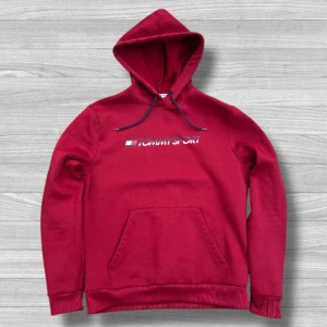 Röd hoodie från Tommy Sport - Snygg röd hoodie från Tommy Sport med logga på bröstet och klassisk känguruficka. Tröjan har huva med mörkblå snören och ribbade muddar. Perfekt för dig som gillar sportig och avslappnad stil.