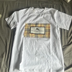 Vit Burberry t-shirt med rutig patch - Vit t-shirt från Burberry i storlek S med klassisk rutig patch framtill och Burberry-logga. T-shirten har rund hals och korta ärmar. Materialet är mjuk bomull och passformen är normal. Perfekt för dig som gillar stilrena och ikoniska designer.