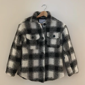 Rutig teddyjacka från Skill, svart/vit - Mysig teddyjacka från Skill i svart och vitt rutigt mönster. Jackan har två bröstfickor med knappar, klassisk krage och stora knappar framtill. Perfekt för dig som vill ha en varm och trendig look med streetkänsla.