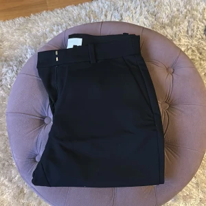 Svarta kostymbyxor från H&M, stl 38 - Klassiska svarta kostymbyxor från H&M i storlek 38. Byxorna har hällor för bälte, två bakfickor och stängs med hyska och hake. Materialet är en mix av polyester, viskos och elastan för en bekväm passform.