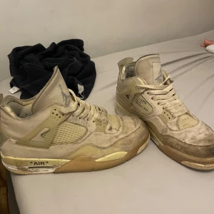 Nike Air Jordan 4 Off-White Sail - Exklusiva Nike Air Jordan 4 Off-White i färgen Sail. Beige och krämvit färgskala med mesh och skinn, ikoniska detaljer som 'AIR'-text på sulan och Off-White branding. Hög modell med snörning och genomskinliga paneler för en unik look.