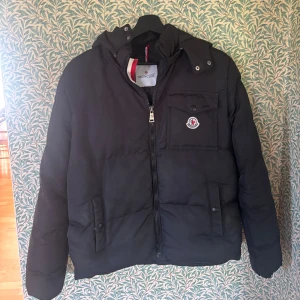 Moncler jacka - Svart pufferjacka från Moncler med huva, dragkedja framtill och flera fickor med tryckknappar. Klassisk Moncler-logga på bröstfickan och snygga detaljer i rött och vitt på insidan av kragen. Perfekt för kalla dagar och riktigt stilren. Om ni har några frågor är det bara att skicka ett meddelande. Storlek 16 men sitter som en small