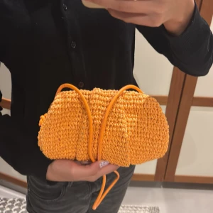 Orange clutch väska från ARKET - Säljer en superfin orange clutch väska från ARKET. Väskan är virkad med mjukt material och har en rundad form. Den har smala axelband och ett snyggt innerfoder i matchande orange tyg. Perfekt för att liva upp din outfit med en färgklick! (Nypris 1225 kr)
