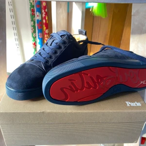 Mörkblåa sneakers från Louboutin - Snygga mörkblå sneakers från Christian Louboutin i mocka med klassisk röd sula och broderad logga på hälen. Skorna har rund tå, snörning och diskreta detaljer som ger en lyxig känsla. Perfekta för dig som vill sticka ut med stil