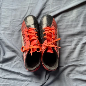 Röd-svarta fotbollsskor från Puma - Säljer ett par fotbollsskor från Puma i rött och svart med coola ränder och silverfärgad logga på sidan. Skorna har snörning och rund tå, med en platt sula som passar perfekt för spel på planen. Materialet är syntet och insidan är svart för extra kontrast.