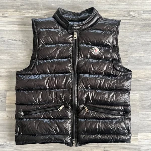 ÄKTA: moncler väst  - Snygg svart dunväst från Moncler med glansig finish och quiltad design. Västen har två dragkedjefickor framtill, hög krage och Moncler-logga på bröstet. Perfekt för lager-på-lager och har en modern, clean look.