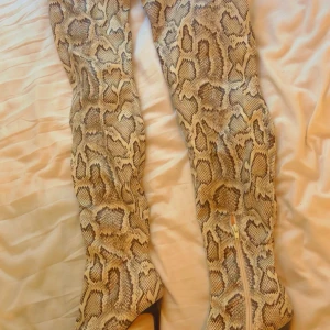 Ormmönstrade höga stövlar med klack - Säljer ett par höga stövlar med ormmönster i beige och bruna toner. Stövlarna har spetsig tå, hög klack och dragkedja på sidan. Perfekta för dig som vill sticka ut med en edgy look.