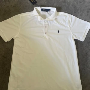 Vit Polo t-shirt från Ralph Lauren - Vit klassisk Polo t-shirt från Ralph Lauren med korta ärmar och krage. T-shirten har tre knappar framtill och den ikoniska broderade loggan på bröstet. Tillverkad i mjuk bomull som känns skön mot huden. Perfekt för en clean och stilren look.