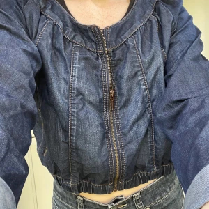 Mörkblå croppad jeansjacka med dragkedja och fickor. - Säljer en mörkblå croppad jeansjacka med dragkedja framtill och elastisk midja. Jackan har långa ärmar, markerade sömmar och fickor på vardera sida. Storleken är large men funkar bra för en oversized look om man är storlek small som jag också.