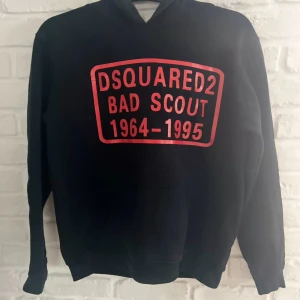 Svart hoodie från Dsquared2 - Svart hoodie från Dsquared2 med stor röd print på bröstet där det står 'DSQUARED2 BAD SCOUT 1964-1995'. Väldigt fin hoodie storleks lappen är borta men passar 165-170 skulle jag säga. 
