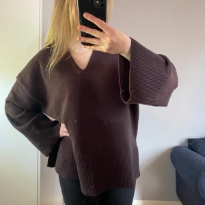 Brun oversized stickad tröja - Mysig oversized stickad tröja i mörkbrunt med v-ringning och breda, långa ärmar. Tröjan har ribbade detaljer vid ärmslut och nederkant samt slitsar i sidorna. Perfekt för lager-på-lager och chill dagar.