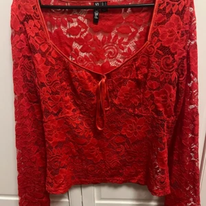 Röd spetsblus från Pieces - Säljer en röd långärmad blus från Pieces i genombruten spets med blommigt mönster. Toppen har v-ringning med knyt framtill och är figurnära. Perfekt för dig som vill sticka ut med en färgstark och feminin look.