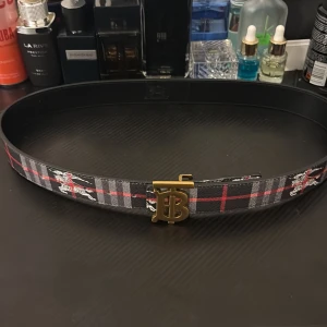 Svart Burberry bälte  - Snyggt bälte från Burberry med klassiskt rutmönster i svart, grått och rött. Bältet har ett stort guldfärgat TB-spänne och detaljer med Burberrys ikoniska riddare. Priset kan diskuteras