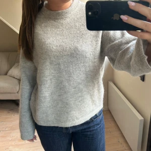 Grå stickad tröja från Gina Tricot - Mysig grå stickad tröja från Gina Tricot med rund halsringning och ribbade muddar. Tröjan har en enkel och stilren design, perfekt att matcha med jeans eller kjol. Materialet känns mjukt och värmande, och passformen är normal med långa ärmar.