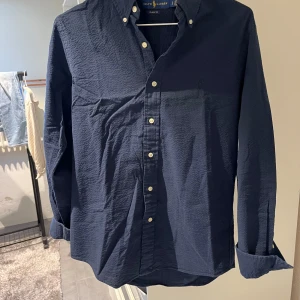 Mörkblå skjorta från Ralph Lauren - Säljer en klassisk mörkblå skjorta från Ralph Lauren i 100% bomull. Skjortan har lång ärm, knappar framtill och en stilren krage. Perfekt till jeans eller chinos för en clean look. Skön och lätt struktur i tyget som ger en avslappnad känsla.
