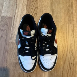 Nike Dunk Low svart/vit/röd - Säljer ett par Nike Dunk Low sneakers i svart, vitt och rött. Skorna har klassisk låg siluett, svart snörning och röd swoosh på sidorna. Ovandelen är i skinn med perforerad tåbox för extra ventilation. Perfekta för dig som gillar streetstyle och vill sticka ut.
