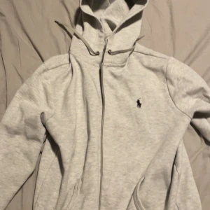 Grå hoodie från Polo Ralph Lauren - Säljer en ljusgrå hoodie från Polo Ralph Lauren med dragkedja och huva. Tröjan har det klassiska broderade logotypen på bröstet och är tillverkad i mjuk bomull. Perfekt för chill dagar eller när du vill ha en clean och stilren look.