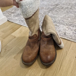 Bruna höga boots i skinn och mocka - Snygga höga boots i brunt skinn med beige mockaskaft och spänndetalj vid sidan. Insidan är fodrad med mjukt vitt foder för extra värme. Rund tå och robust sula gör dem perfekta för kyligare dagar. Perfekt till jeans eller kjol för en cool look.