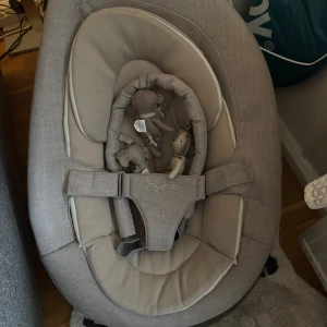 Nuna Leaf Grow Babysitter - Nuna Leaf Grow babysitter i beige/grått tyg. Ergonomisk design, mjuk sits och säkerhetssele. Inkluderar leksaksbåge med mjuka figurer. Tyget ser fräscht ut, inga synliga fläckar eller skador. Perfekt för småbarn, ger naturlig gungning utan batterier.