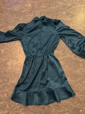 Mörkblå satin klänning med volang - Supersnygg mörkblå klänning i satin med lång ärm och hög krage. Klänningen har markerad midja, volang nertill och en lätt glansig finish som ger en lyxig känsla. Perfekt för dig som vill sticka ut med stilren elegans.