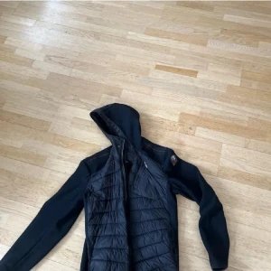 Svart quiltad pufferjacka med huva - Svart pufferjacka från Blåkläder med quiltad framsida och rygg, huva och dragkedja. Jackan har orange detaljer på insidan och flera praktiska fickor. Materialmix med polyester och syntet, perfekt för kyliga dagar. Snygg och funktionell design för dig som gillar streetwear.