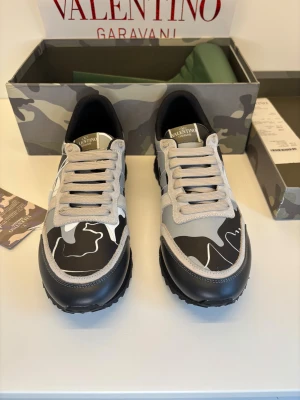 Valentino Garavani Rockrunner – Storlek 42 – Grå/Svart Camo – Toppskick! - Säljer ett par exklusiva Valentino Rockrunner i storlek 42, ikoniska sneakers kända för sin lyxiga design och oslagbara komfort. Modellen kommer i den klassiska camo-looken i grå/svarta toner med stilrena detaljer och de karakteristiska nitarna vid hälen.