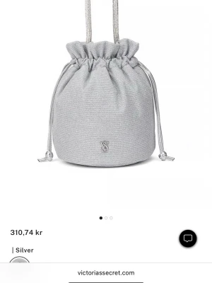 Silver handväska från Victoria's Secret - Glittrig silverfärgad handväska från Victoria's Secret i bucket-modell med dragsko och gnistrande handtag. Väskan har VS-logga framtill och skimrande tyg som ger en lyxig känsla. Perfekt för dig som vill sticka ut med en snygg accessoar.