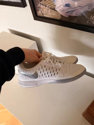 Nike lunargaato  - Nike Hyperpulse fotbollsskor i vitt med silverfärgade detaljer och sula. Skorna har snörning, quiltad ovandel och ventilerande meshpaneler på sidorna. Snygg, sportig design med rund tå och platt sula, perfekt för inomhusspel. Materialet är syntet och skinn.