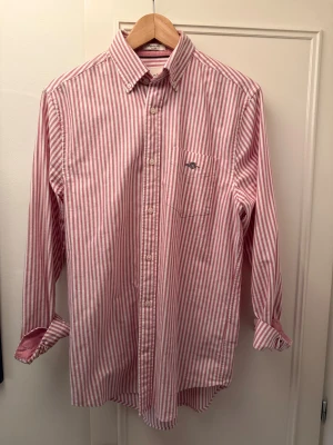 Randig skjorta från Gant i bomull - Säljer en klassisk skjorta från Gant med rosa och vita vertikala ränder. Skjortan har button down-krage, bröstficka med broderad logga och knäppning framtill. Tillverkad i mjuk bomull och har långa ärmar. Perfekt för en preppy look.