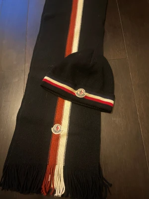 Moncler svart stickad mössa & halsduk - Svart stickad mössa och halsduk från Moncler med klassiska logopatchar. Mössan har röd, vit och marinblå rand vid kanten. Halsduken har ränder i rött, vitt och svart samt fransar i ändarna. Perfekt för att hålla stilen under kalla dagar. Äkta