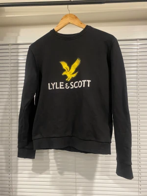 Svart Lyle & Scott sweatshirt S - Svart sweatshirt från Lyle & Scott i storlek S. Tröjan har ett stort gult fågeltryck och vit logga på bröstet. Rund hals, ribbade muddar och lång ärm. Mjuk bomullskvalitet som är skön att ha på sig. Perfekt för dig som gillar klassisk streetstil.