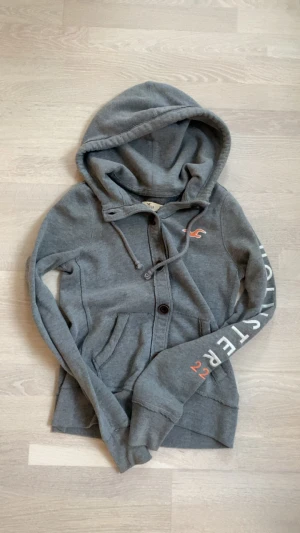 Grå hollister hoodie med knappar - Grå hoodie från Hollister i mjuk bomull med knappar framtill och huva. Långärmad modell med logotyp i orange på bröstet och tryck på ena ärmen. Perfekt för chill dagar och enkel att matcha med jeans eller joggers.