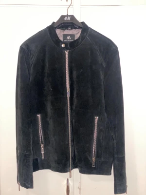 Svart mockajacka från Jofama x Måns Zelmerlöw - Svart mockajacka med coola silverfärgade dragkedjor framtill och vid fickorna. Jackan har en stilren, rak passform och är helsvart med mjuk mockayta. Perfekt för dig som vill ha en snygg och tidlös look med edge.