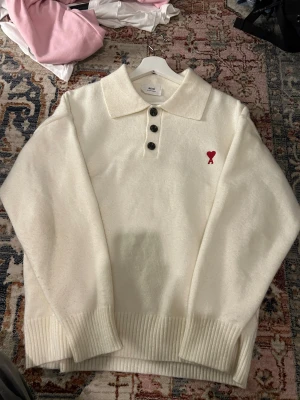 Vit stickad pikétröja från Ami - Vit stickad pikétröja från Ami med klassisk krage och tre svarta knappar framtill. På bröstet finns Ami:s ikoniska röda hjärta med A-broderi. Tröjan har ribbade muddar och är långärmad, perfekt för en clean och chill look.